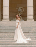 Woman wearing Monique Lhuillier Fall 2023 Bliss white off-the-shoulder crepe and mikado fitted BL23204 gown with detachable bow train