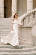 Woman wearing Monique Lhuillier Fall 2023 Bliss white off-the-shoulder crepe and mikado fitted BL23204 gown with detachable bow train