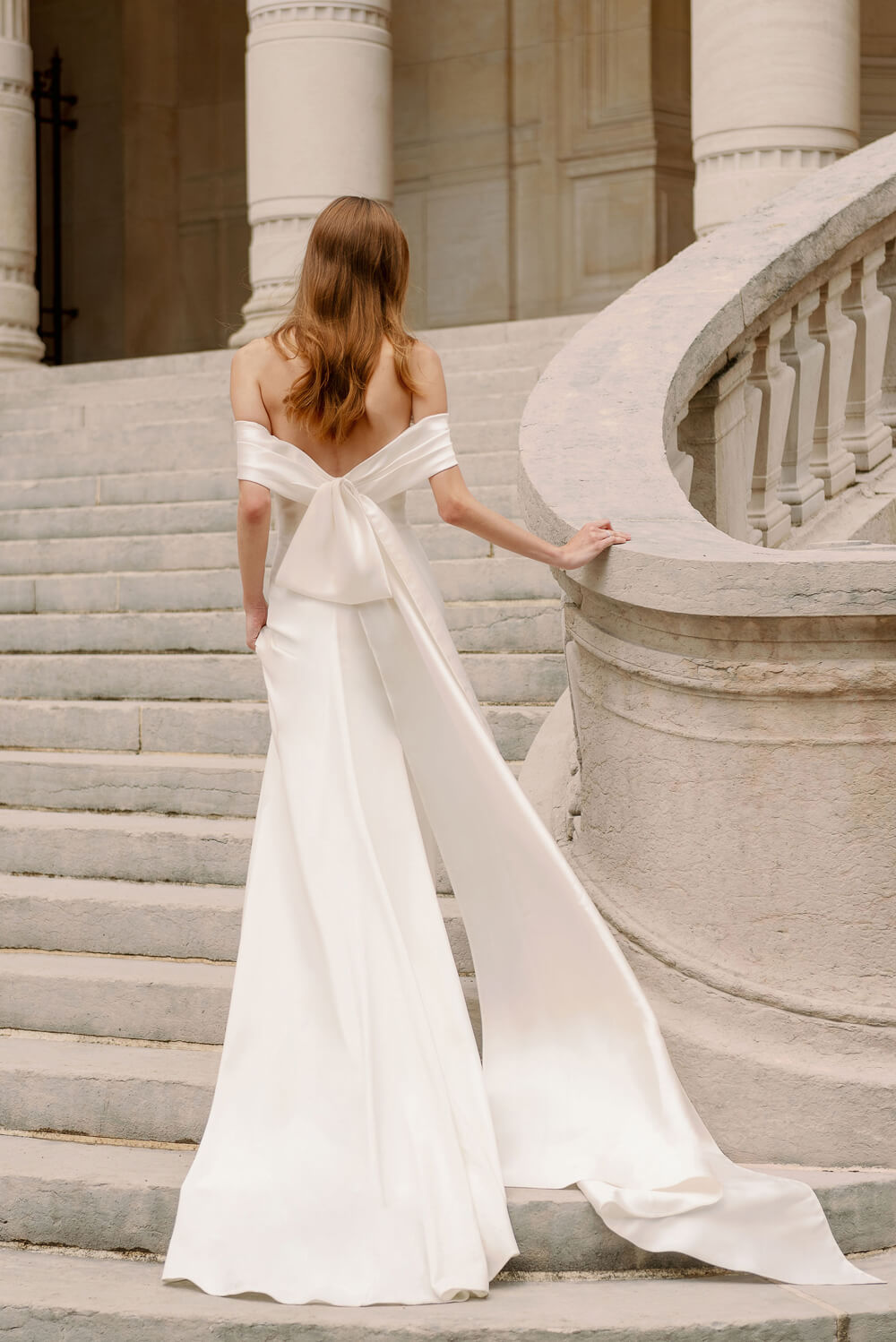 Woman wearing Monique Lhuillier Fall 2023 Bliss white off-the-shoulder crepe and mikado fitted BL23204 gown with detachable bow train