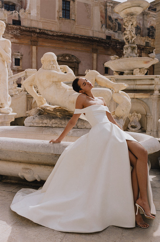 Woman wearing Monique Lhuillier Spring 2023 Bliss white off-the-shoulder mikado a-line BL23109 gown with side slit in skirt