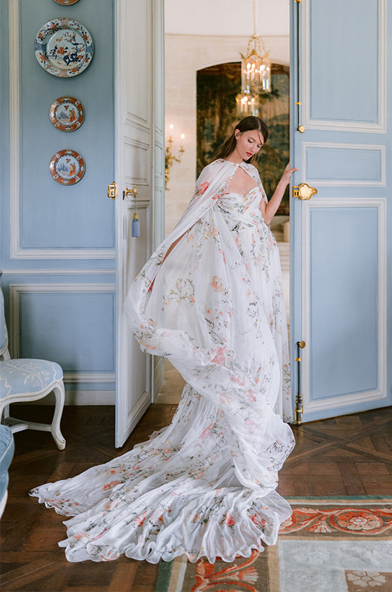 Woman wearing Monique Lhuillier Fall 2023 white and floral printed Bouquet chiffon sheath gown and matching long cape