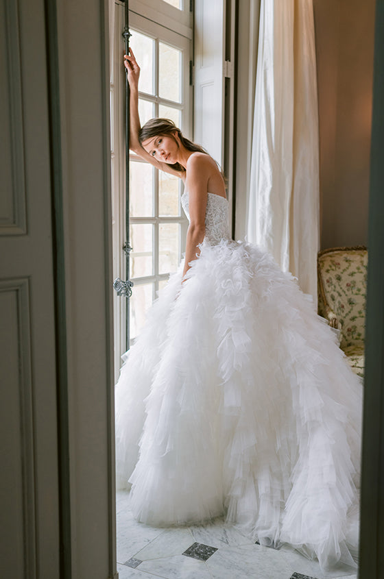 Woman wearing Monique Lhuillier Fall 2023 Birdie white ballgown with embroidered beaded bodice and feather and pleated tulle layered skirt