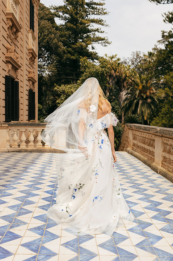 Woman wearing white and multi colored 3D floral embroidered Monique Lhuillier Spring 2023 Liliana off-the-shoulder a-line gown with veil