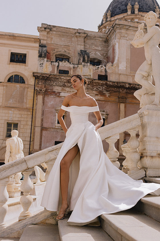 Woman wearing Monique Lhuillier Spring 2023 Bliss white off-the-shoulder mikado a-line BL23109 gown with side slit in skirt