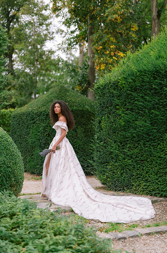 Woman wearing Monique Lhuillier Fall 2023 white and purple off-the-shoulder Lavender floral printed a-line gown