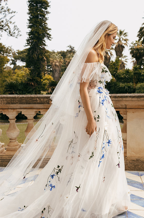 Woman wearing white and multi colored 3D floral embroidered Monique Lhuillier Spring 2023 Liliana off-the-shoulder a-line gown with veil