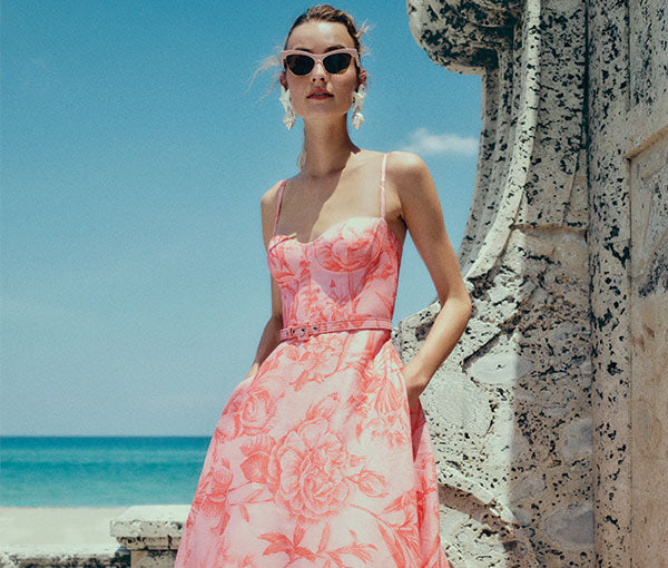 Monique Lhuillier Spring 2025 coral floral linen spaghetti strap a-line dress on model in front of stone wall on beach