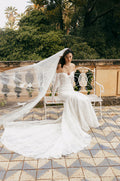 Woman wearing white long sleeve off-the-shoulder mosaic lace Monique Lhuillier Spring 2023 Viviana trumpet gown